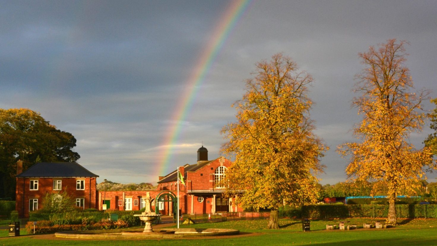 A rainbow above a grassy park in autumn. There is a warm golden light and orange leaves on the trees.