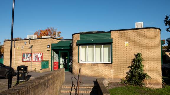 A beige coloured brick community building with dark green entrance and window shutters. There are two notice boards on the wall outside. Steps with a handrail lead up to the building. It's a sunny day with blue skies above.