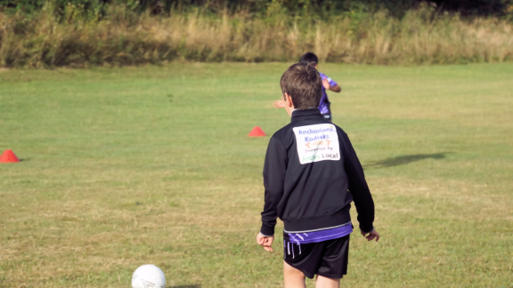 A child in a football kit walks towards a football on a grassy field during an outdoor football training session.