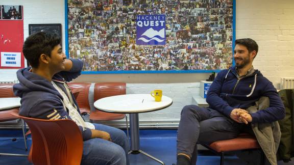 Two young men sit opposite each other in brown chairs at a youth centre.