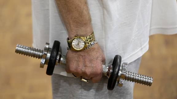 A close-up of a person holding a dumbbell during an indoor exercise activity. They have a gold watch on their wrist.