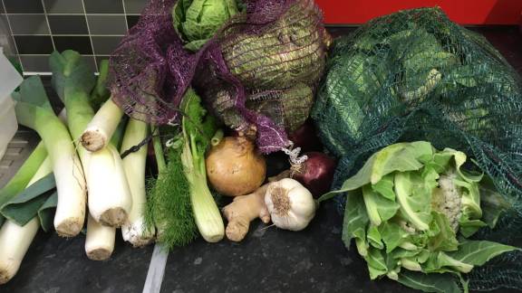 Pile of different vegetables on a grey kitchen counter, including leeks, cabbage and garlic.