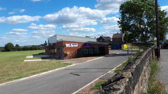 A red brick building on a playing field on a sunny day. In front is a tarmac road and a low stone wall.