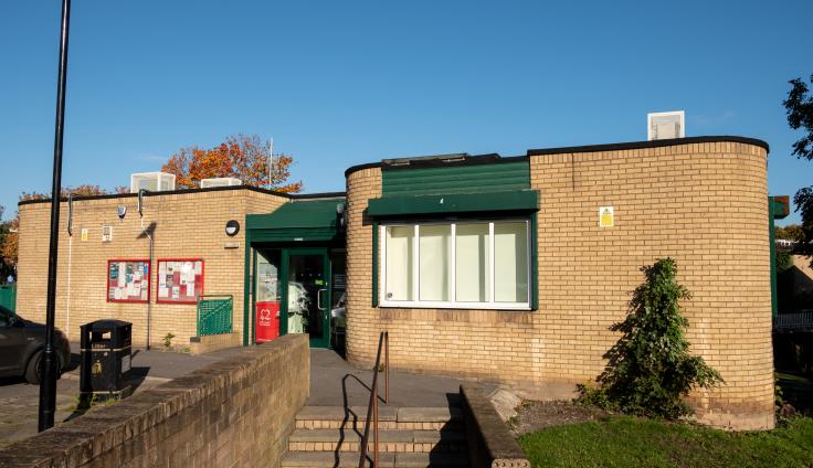 A beige coloured brick community building with dark green entrance and window shutters. There are two notice boards on the wall outside. Steps with a handrail lead up to the building. It's a sunny day with blue skies above.
