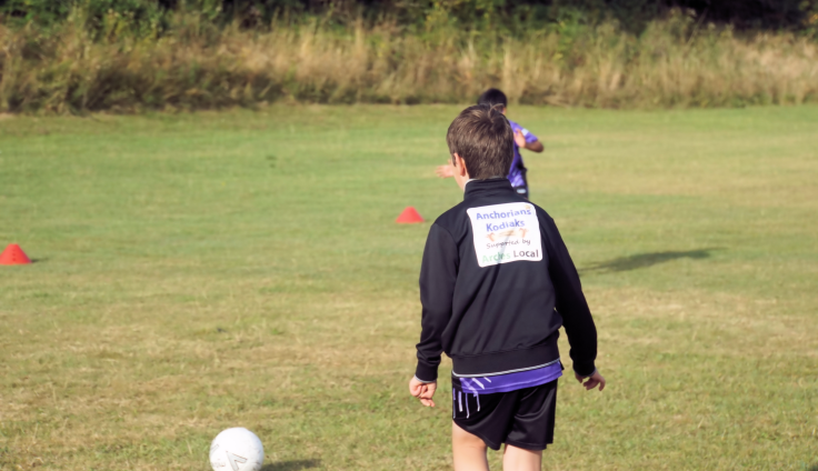 A child in a football kit walks towards a football on a grassy field during an outdoor football training session.