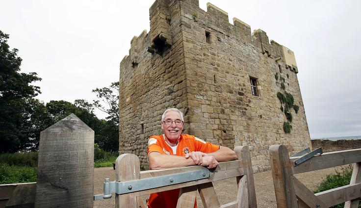 A older man wearing an orange high vis jacket and glasses leans on a wooden fence and smiles. Behind him is a large turreted building.