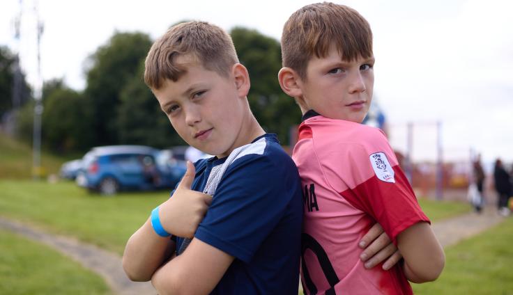 Two local boys stand back to back, one in a blue shirt and one in a pink shirt, looking at the camera, one giving a thumbs up.