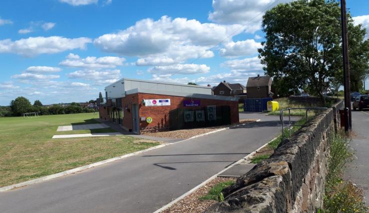 A red brick building on a playing field on a sunny day. In front is a tarmac road and a low stone wall.