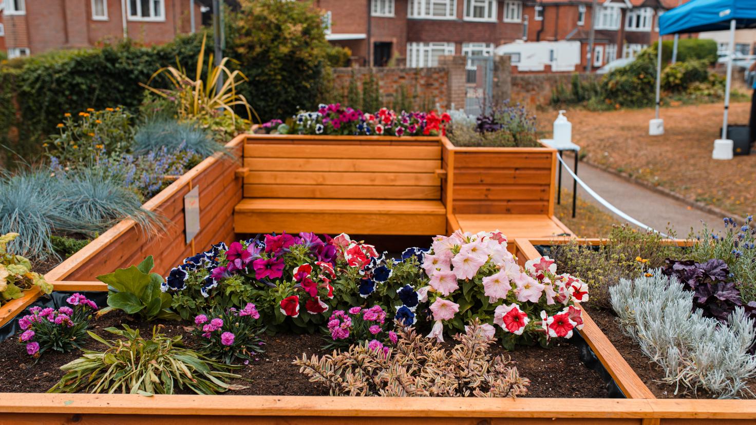 A wooden seating area, planted with flowers, with houses in the distance.