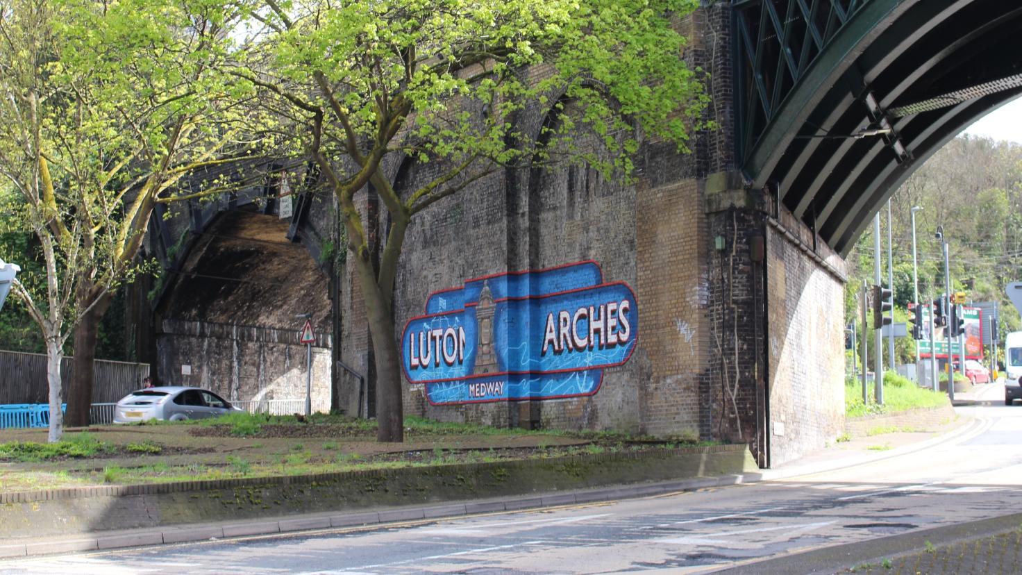 An arched railway bridge with a mural of the words 'Luton Arches' on the adjacent brick wall.