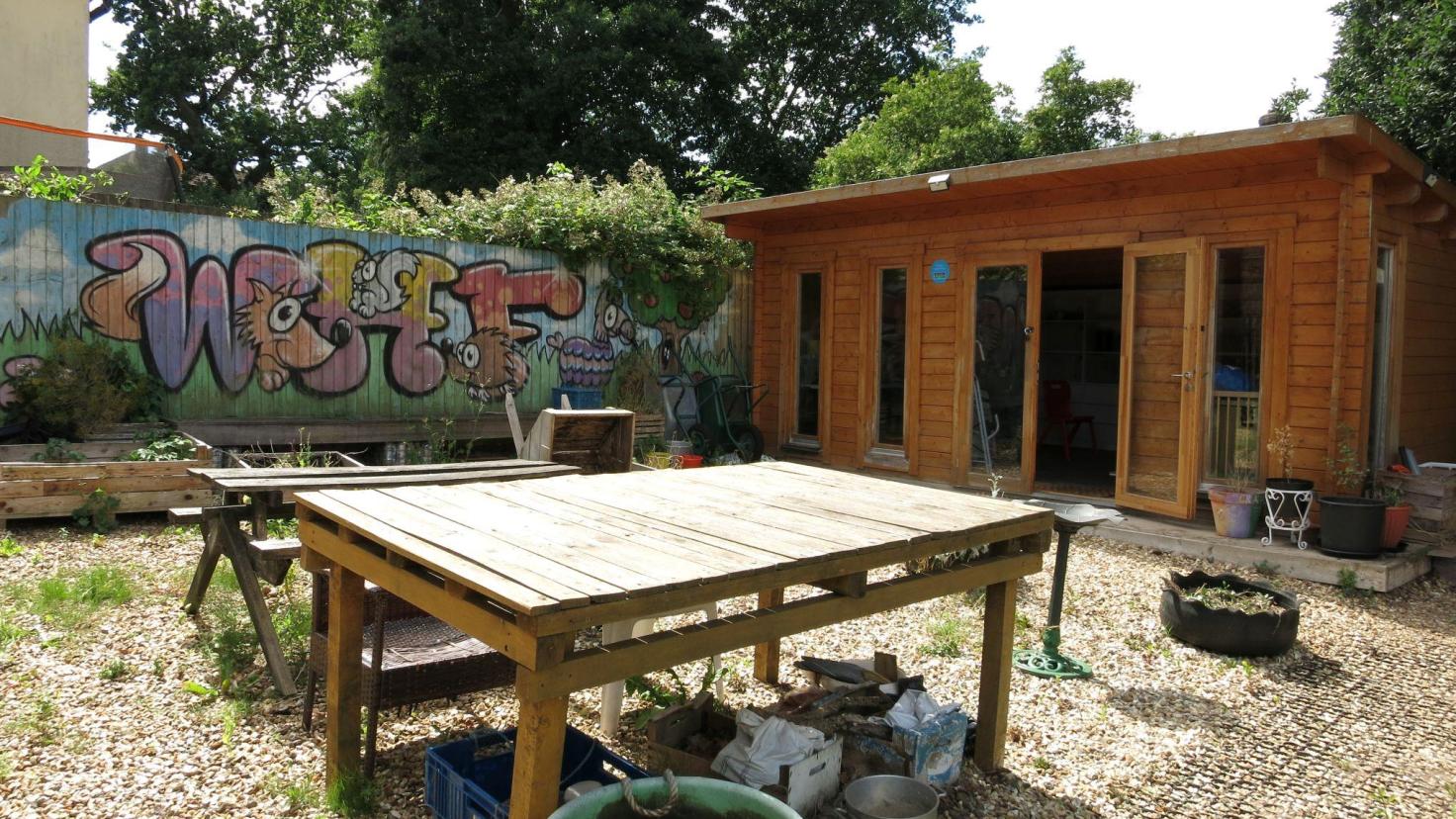 A wooden table in the sunshine in front of a wooden building, with a colourful spray-painted mural on a wall in the background.