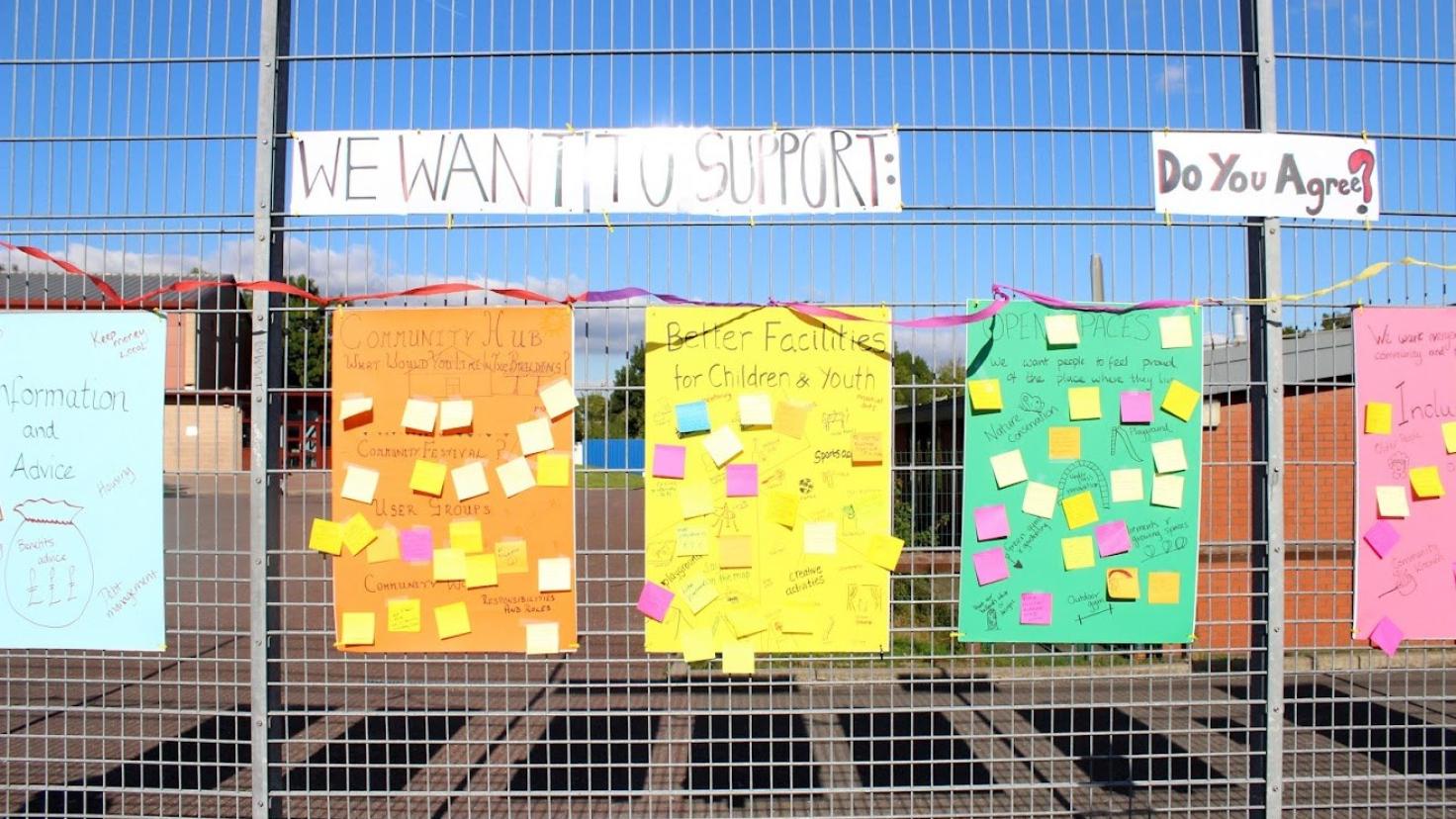 Brightly-coloured posters covered in post-in notes, attached to a steel mesh fence.