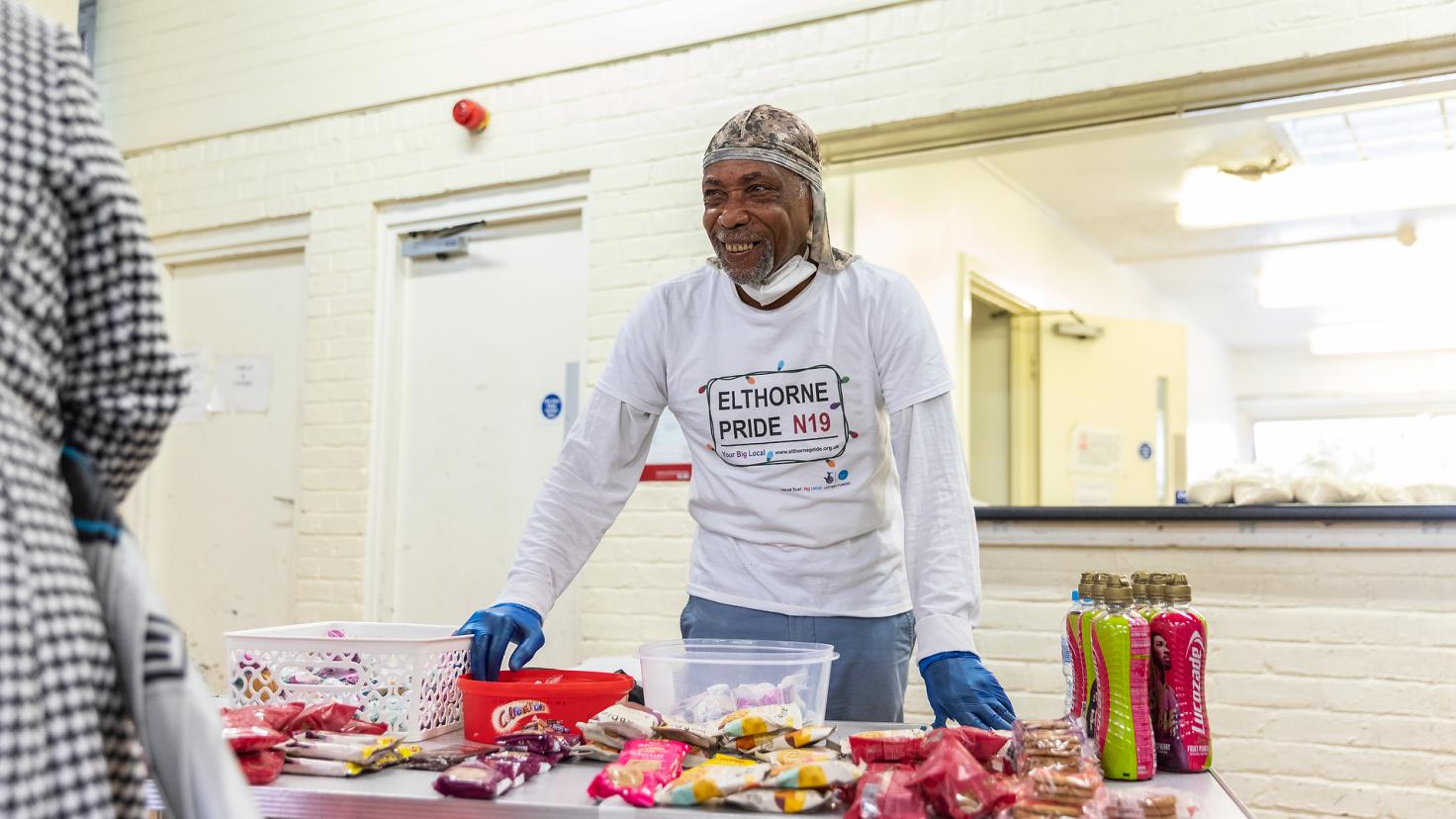 A man wears an apron reading 'I heart Elthorne Pride N19' with food on the table.