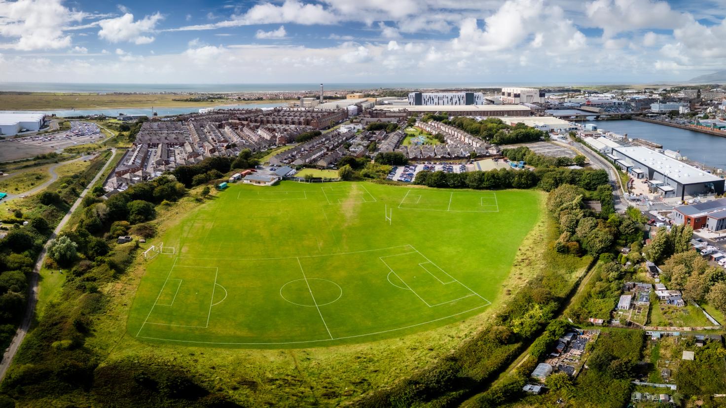 Aerial view of a large green space with football pitch markings in the foreground and buildings in the background and a water channel on the right.