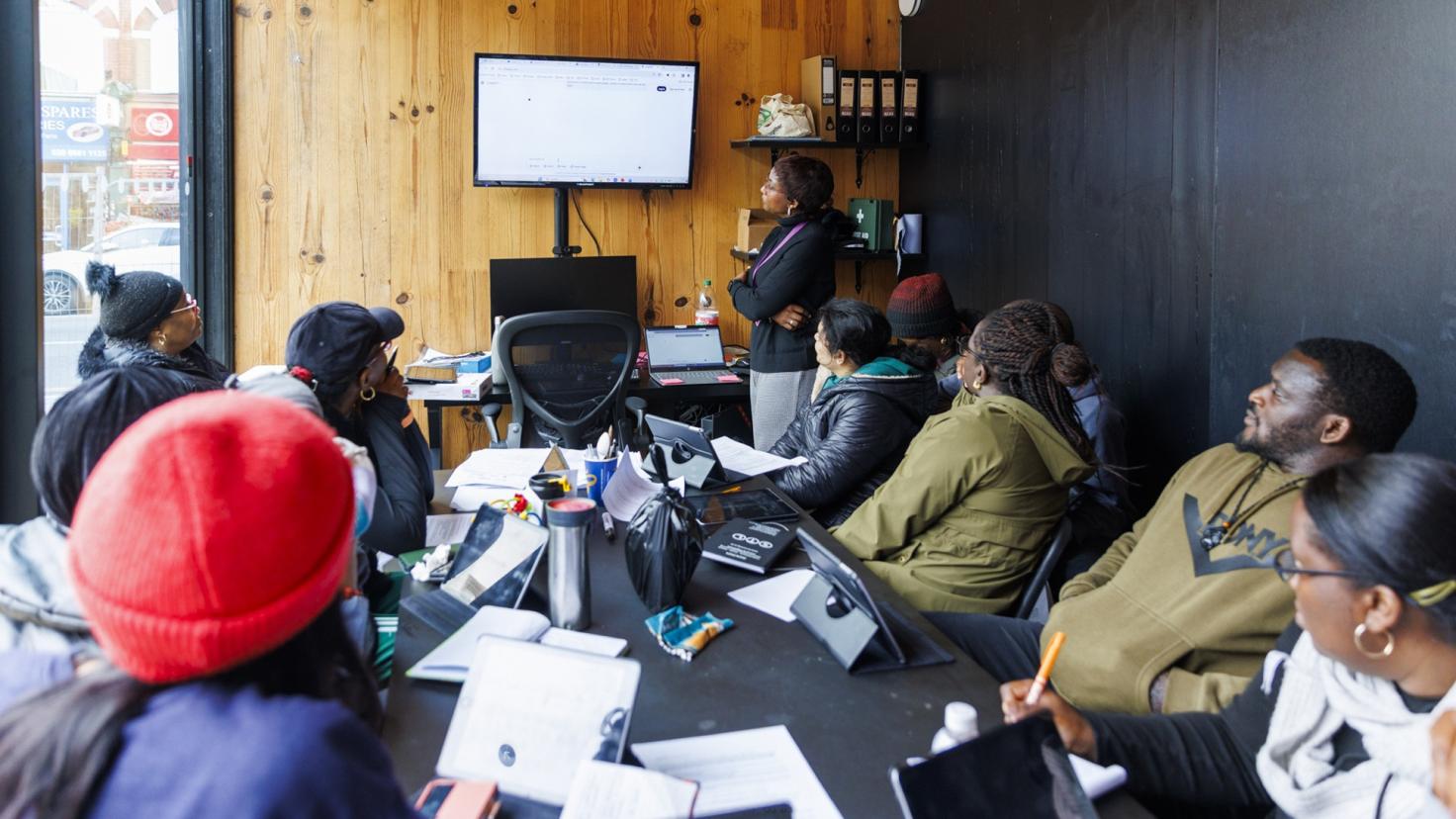 A group of people watching a woman who is facing a presentation screen at the front of the class.