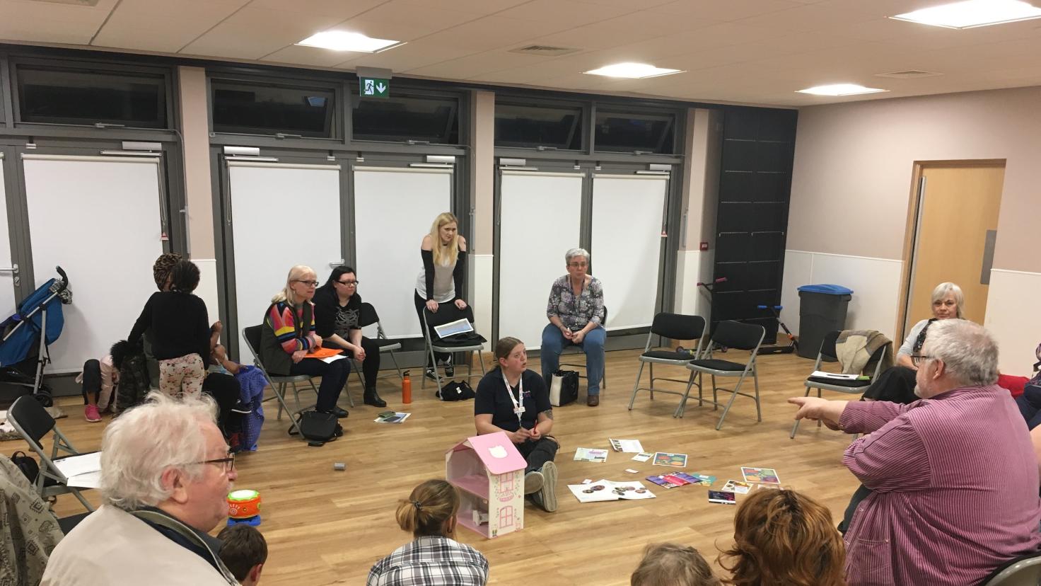 A multi-generational group gathers in a modern community room with large windows and wood floors. Adults and children sit in a loose circle on black folding chairs, with some participants sitting on the floor. A blonde woman in a black sleeveless top sits in the centre. Scattered on the wooden floor are books, papers, and a pink dollhouse.