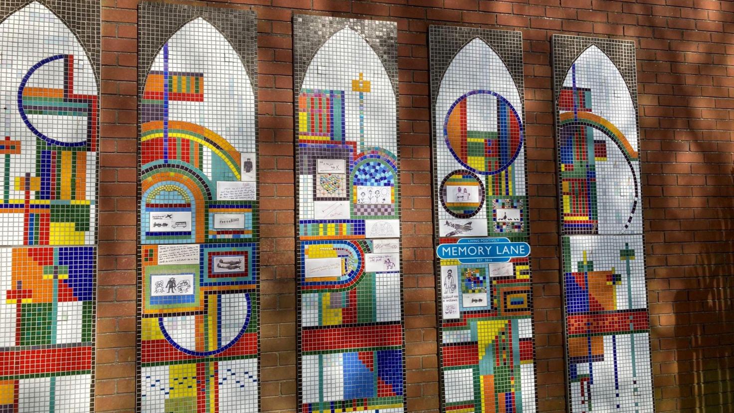 Four colourful mosaics in the shape of arched windows, on a red brick wall.