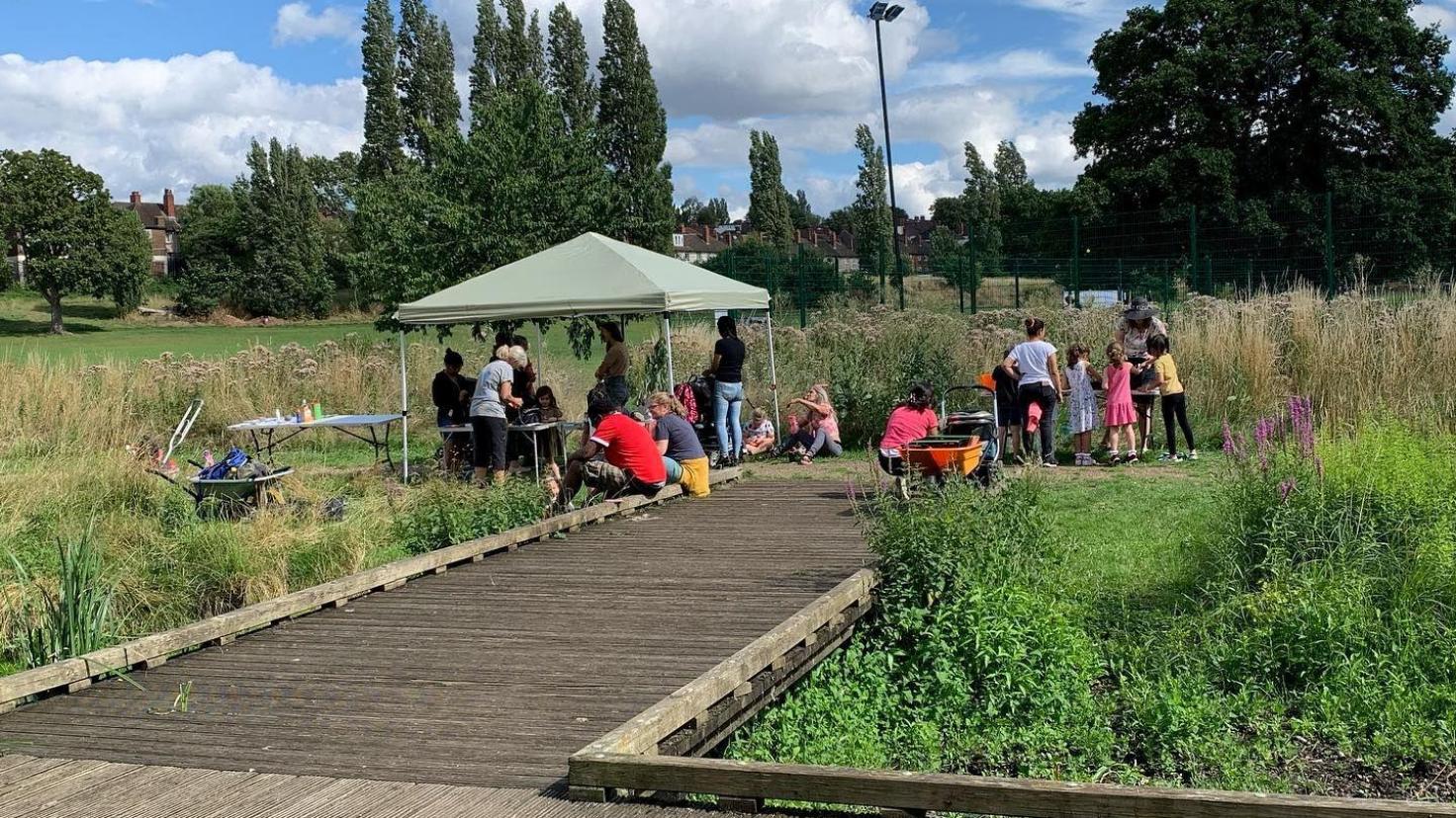 Green gazebo on a wooden jetty with twenty people in a green space.