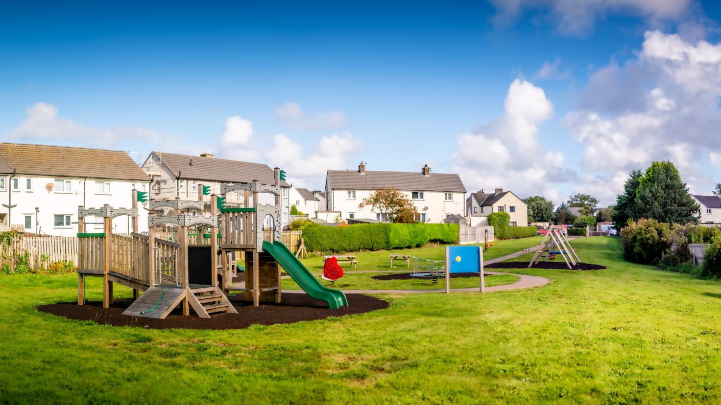 Wooden playground equipment including a slide in a green area behind houses.