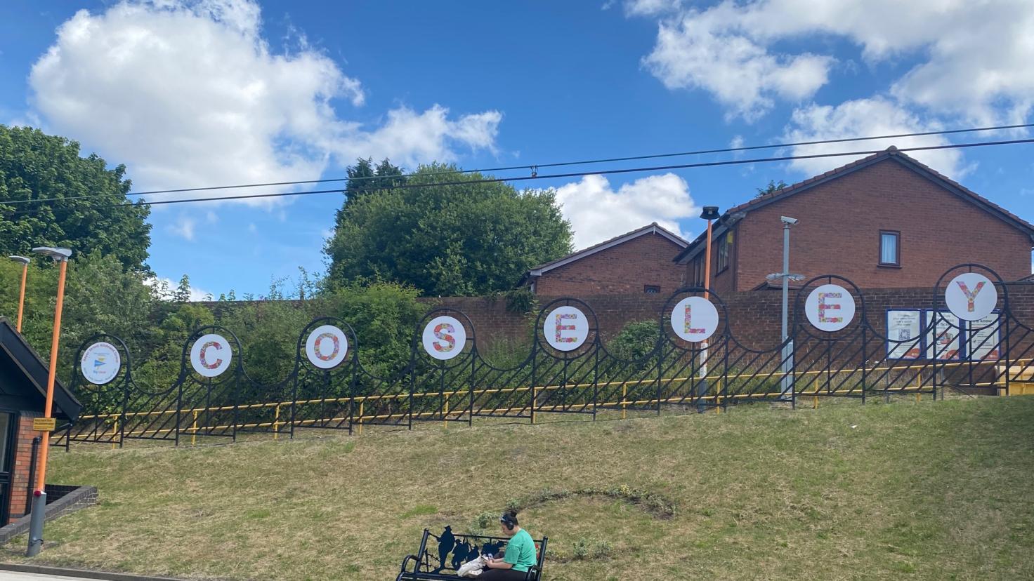 A person sitting on a bench on a railway platform, with letters spelling 'Coseley' on a fence in the background.