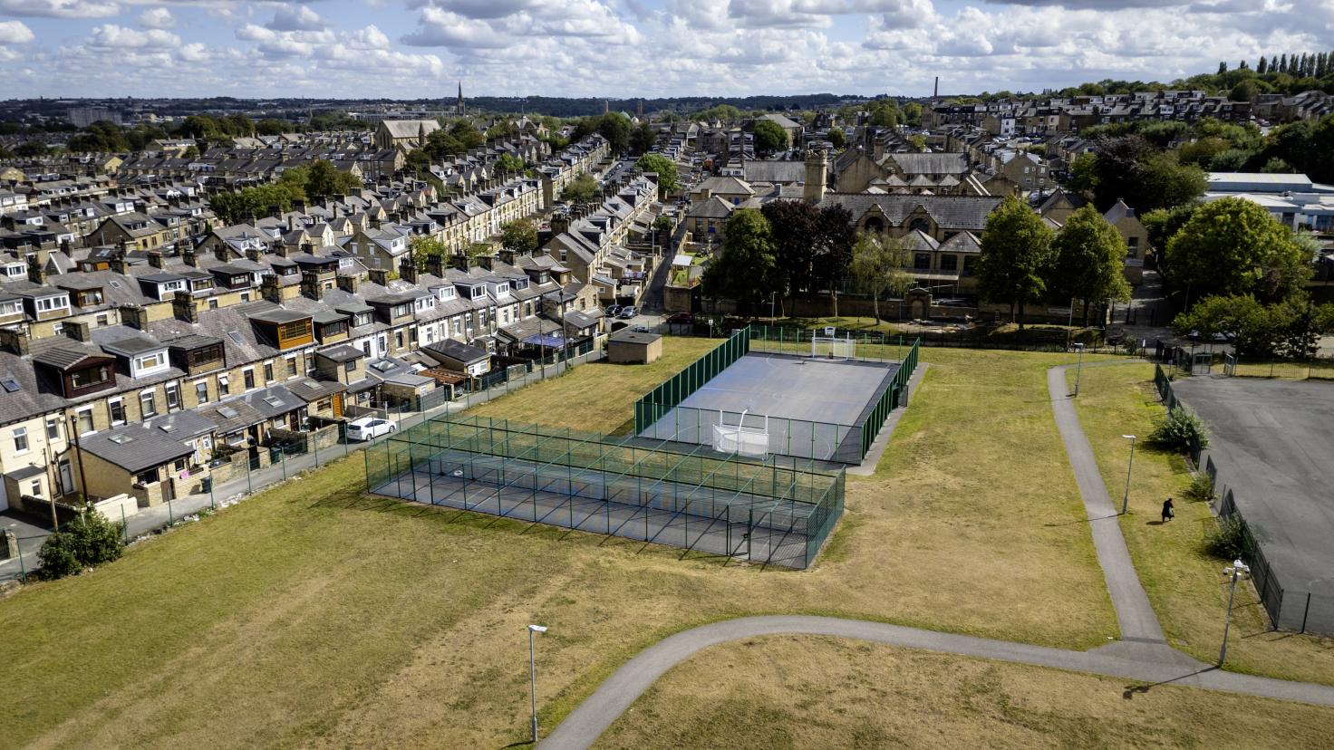 Aerial view of tarmacked sports area surrounded by green fencing, with green space in the foreground and residential houses on the left