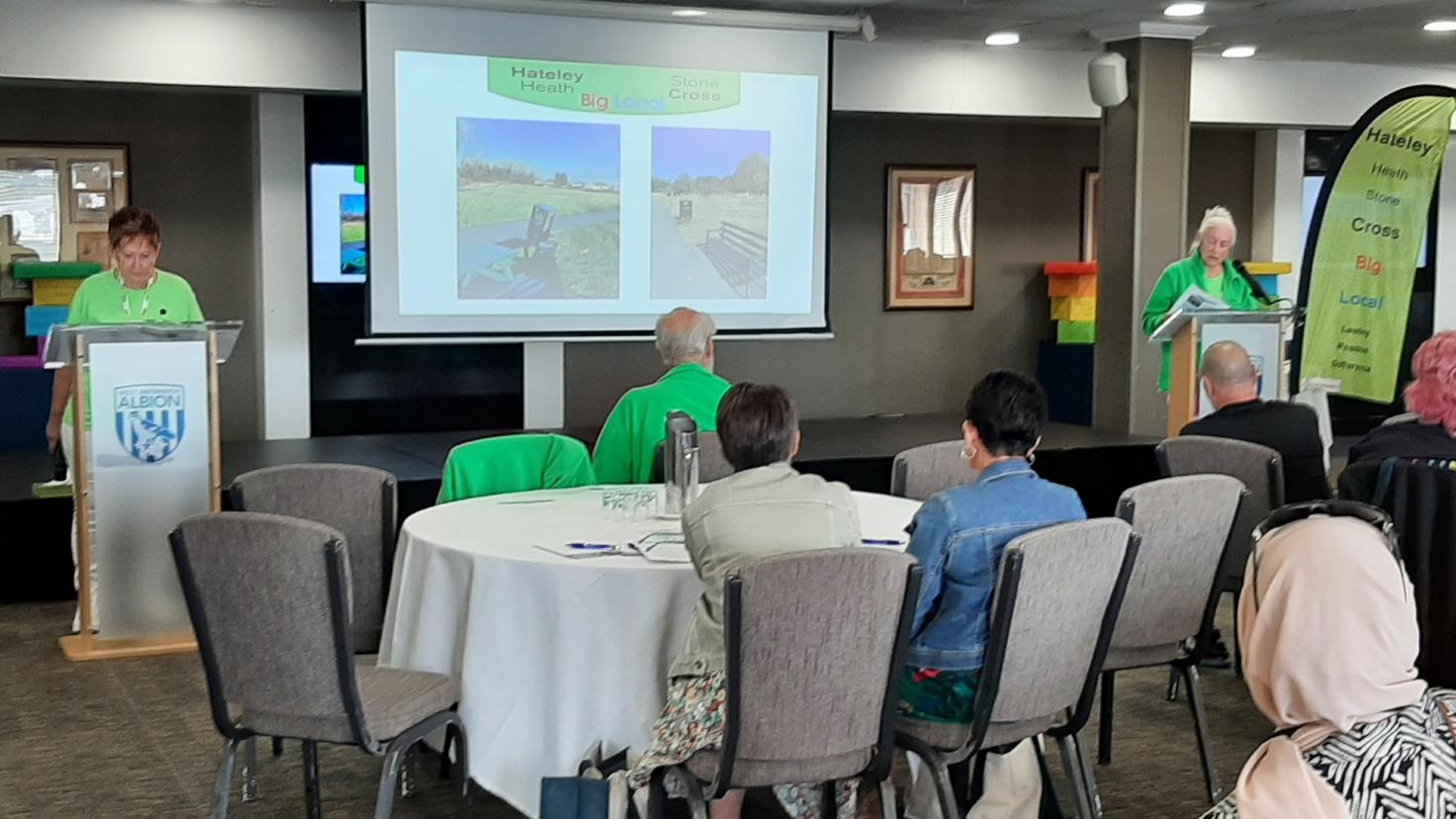People sitting on chairs in a conference room with speakers talking at lecterns and an overhead projector screen.