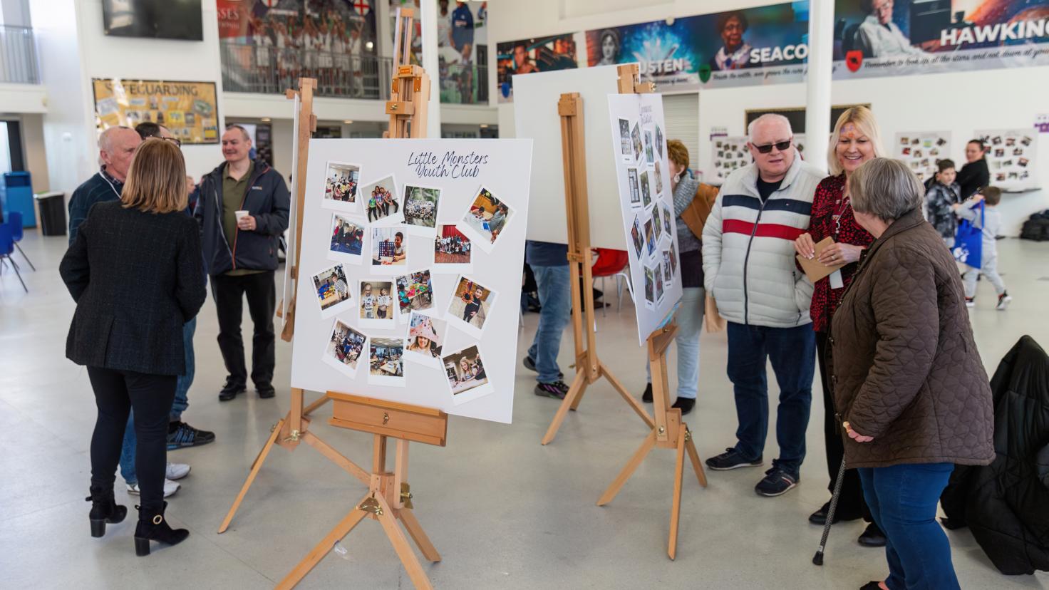 Groups of people stand around talking, surrounded by white boards with photos on them.