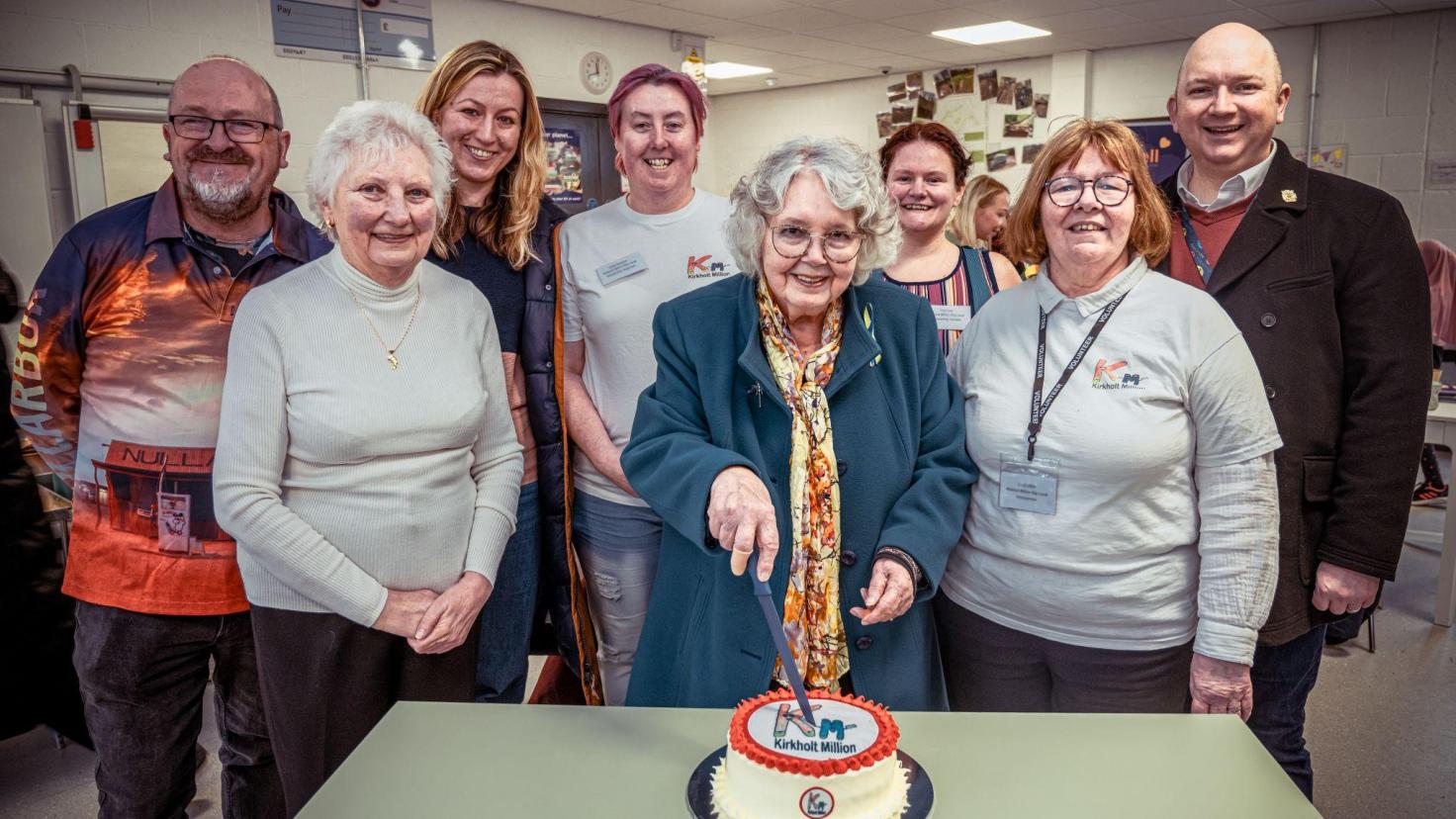 A group of people smiling at the camera, with one person cutting into a cake.