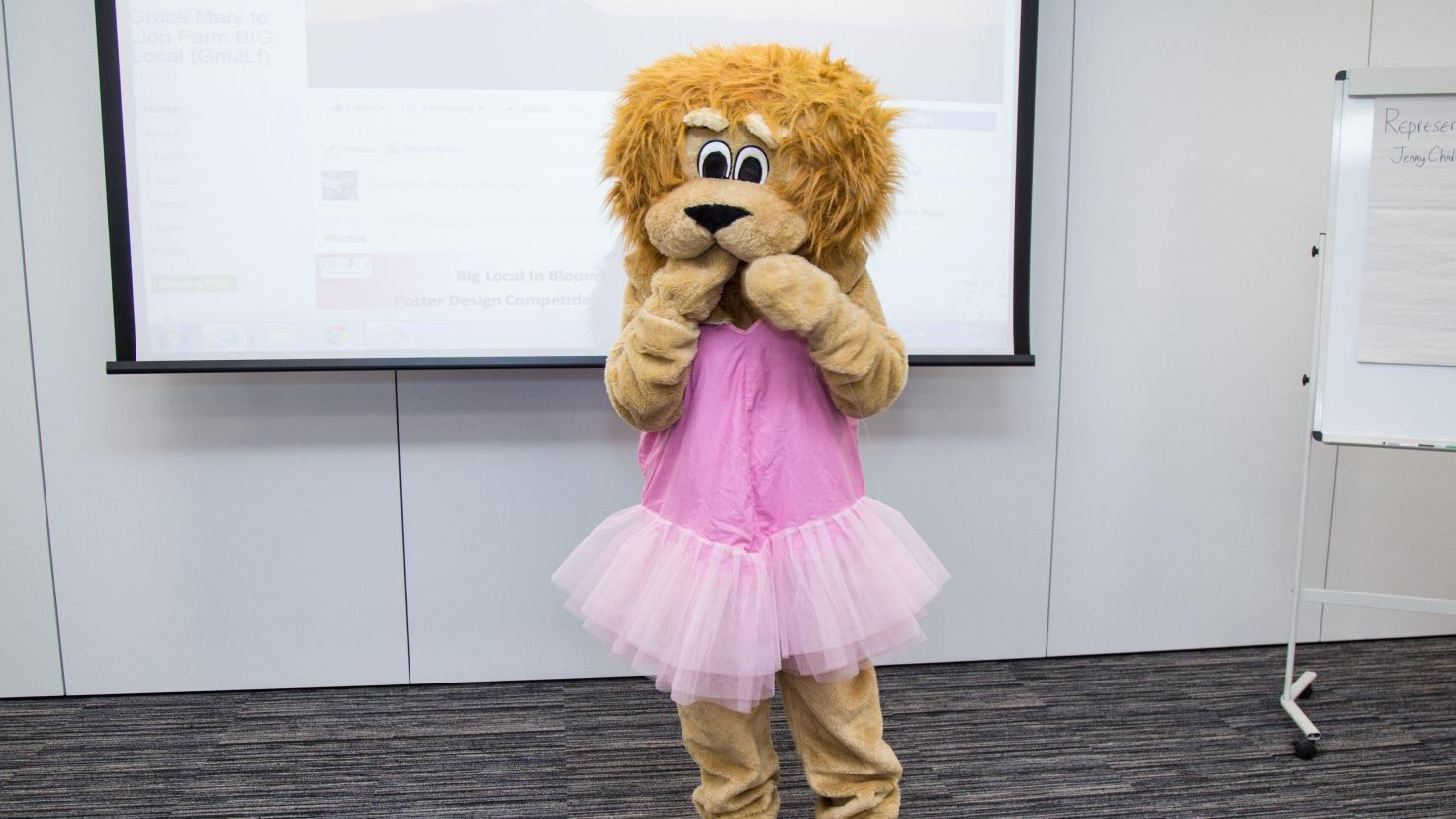 A person wearing a lion costume and a pink tutu standing in front of a screen.