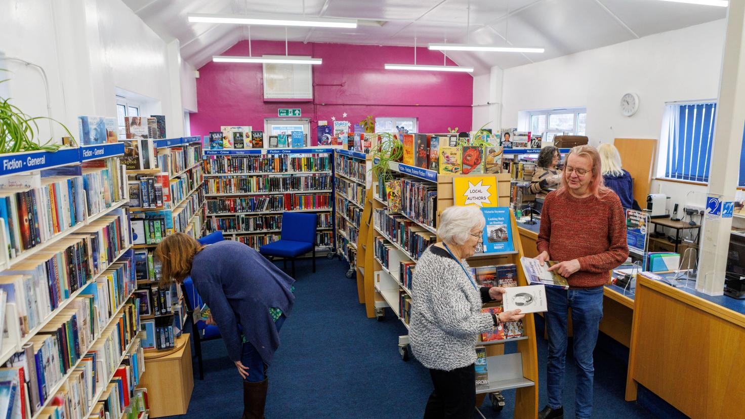 People browsing in a library which has a cerise pink feature wall at the far end.
