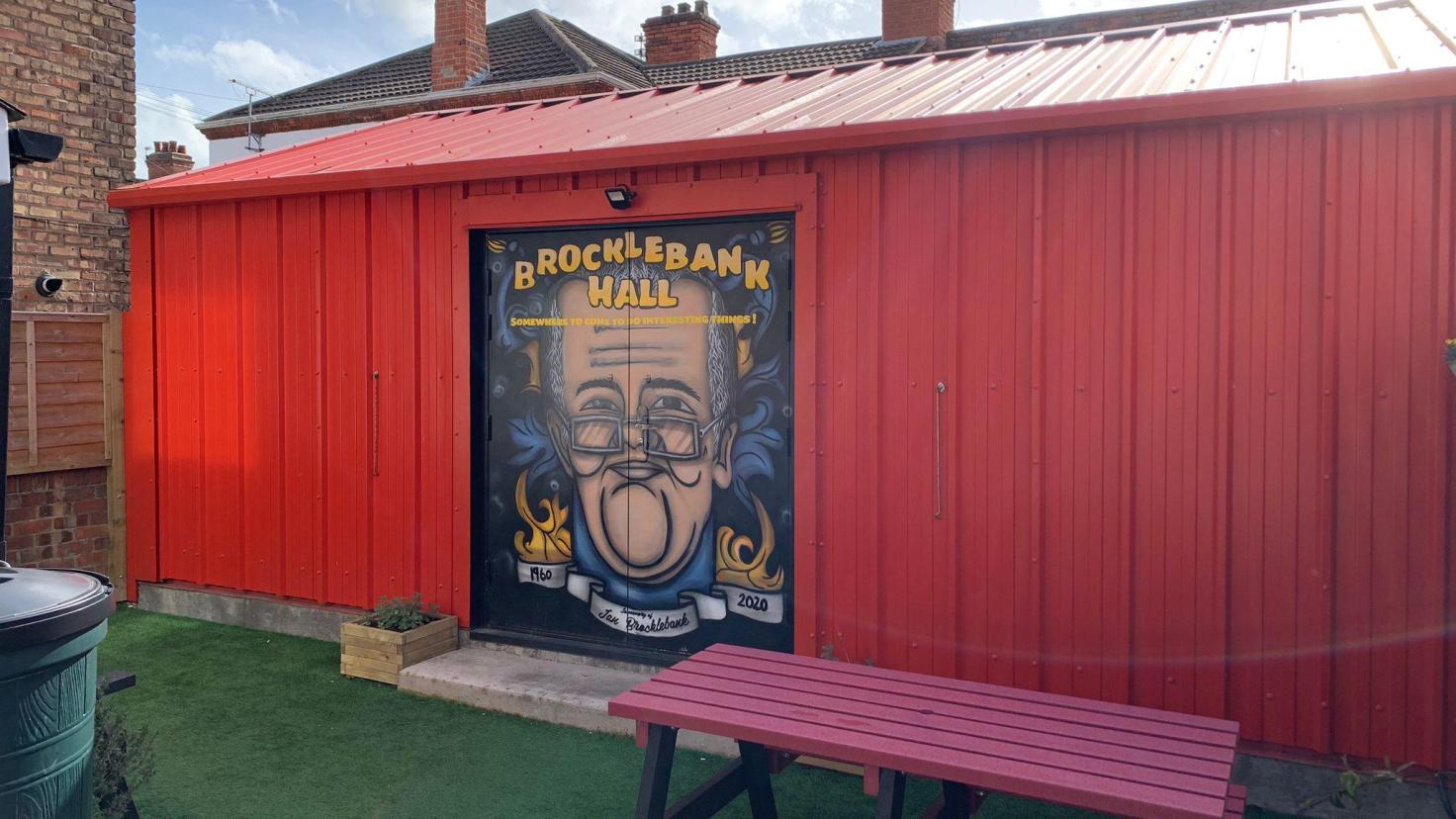 A red shed with 'Brocklebank Hall' painted on the doors along with an image of Ian Brocklebank.