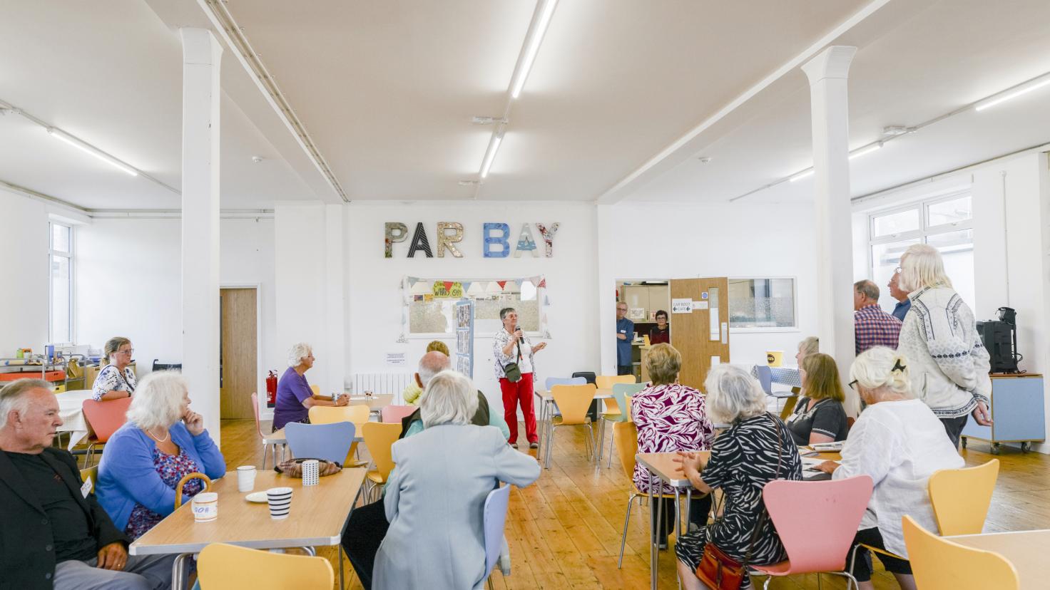 A group of people listening to a woman speak in a community hub.