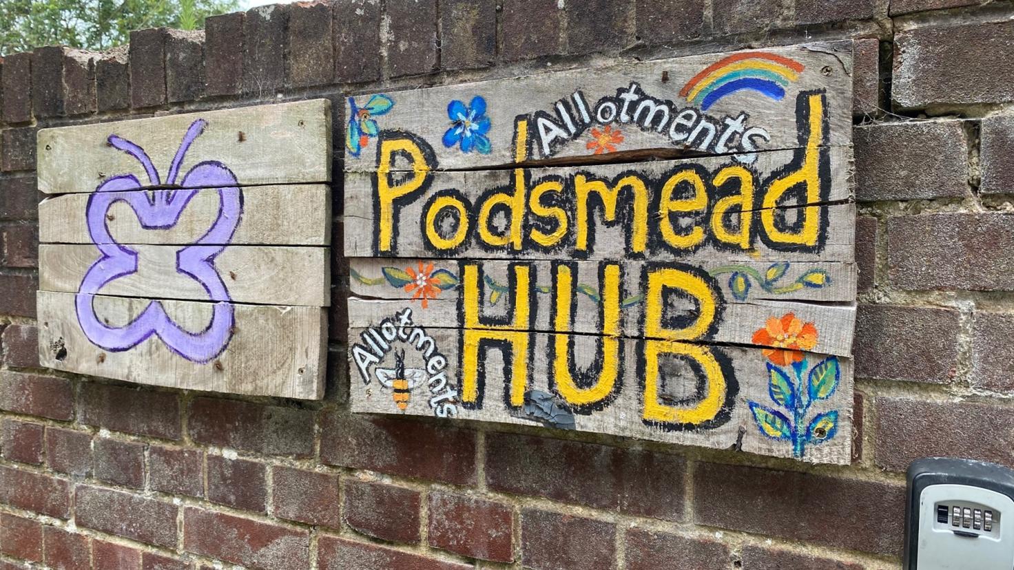 Two recycled palette signs on a brick wall: one with a butterfly outline and one reading 'Podsmead Hub'.
