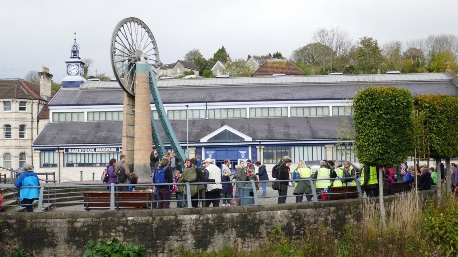 A crowd of people outside, gathering around a brick tower with a large bicycle-wheel type construction on the top.