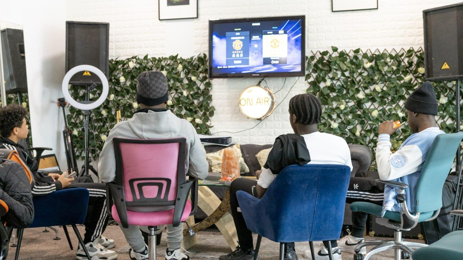 Four people in a room with chairs, facing a monitor, surrounded by tall sound speakers.