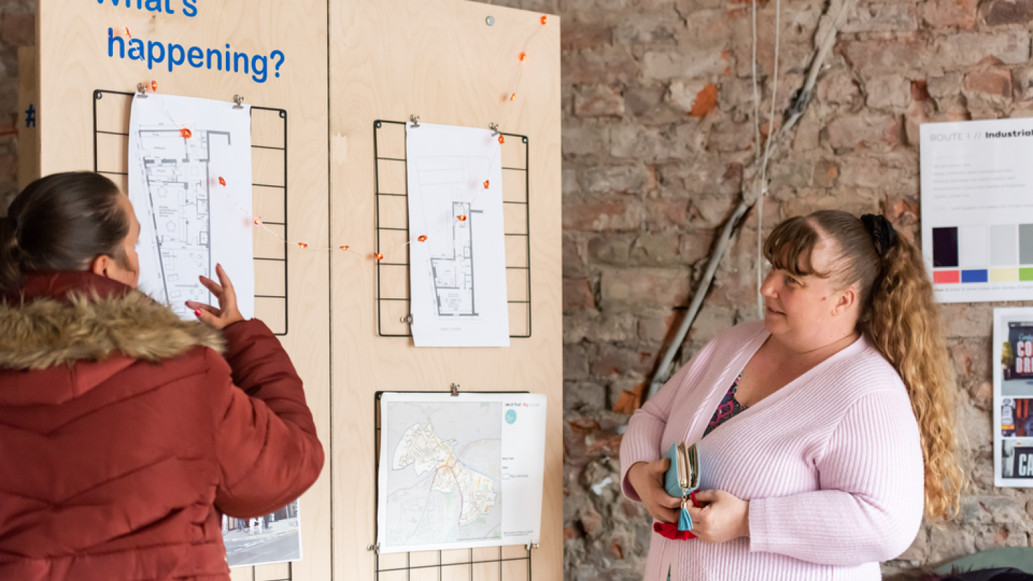Two people are standing next to a wooden board with plans for the community hub pinned to it. Blue text reads 'What's happening?'. They appear to be discussing what's on the board.