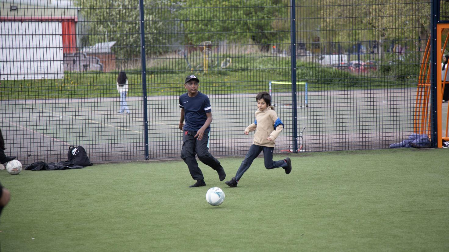 Two people kicking a football in a fenced-off sports area.