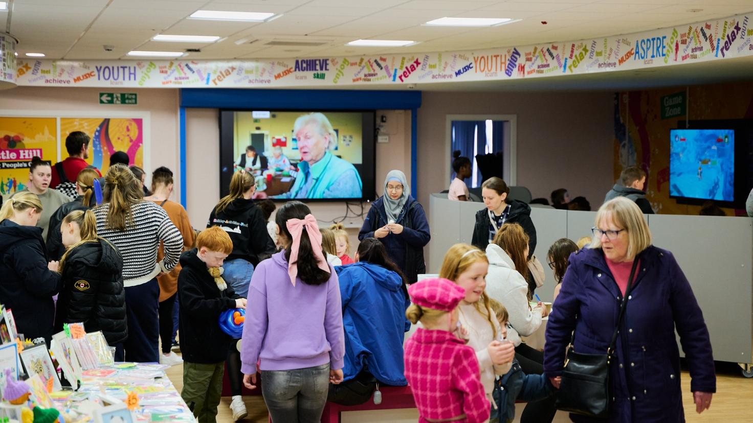 A gathering of people in a large room, watching a television screen next to tables with activities on them.