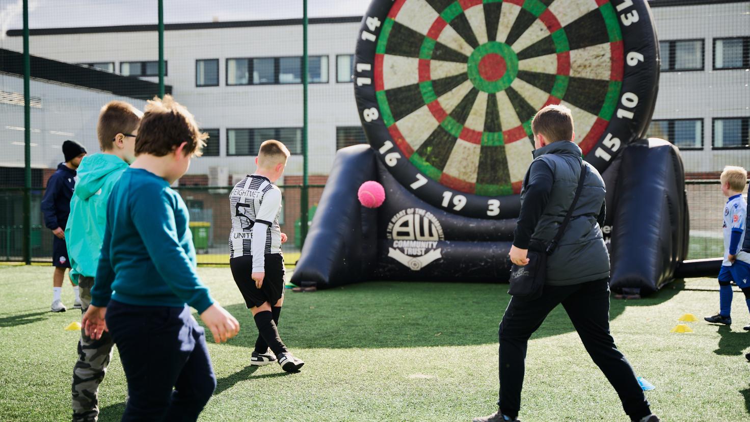 A group of children kicking a football towards a goal, which is a giant inflatable dart board.