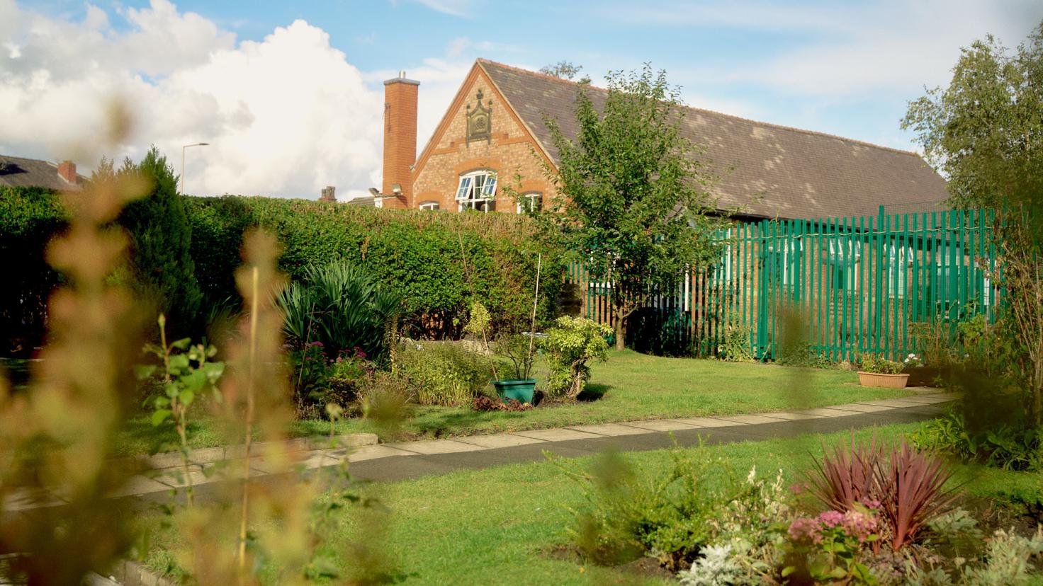 An enclosed green space with a grey path through the centre, green metal fencing and hedge