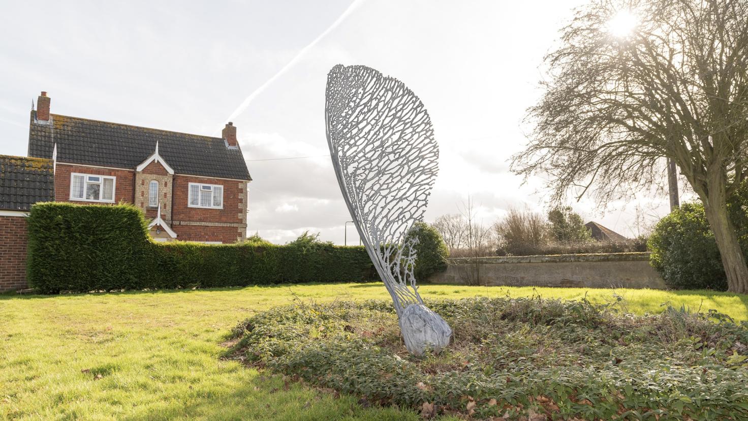 A large metalwork sculpture of a sycamore seed in a grassy area with a house in the background.