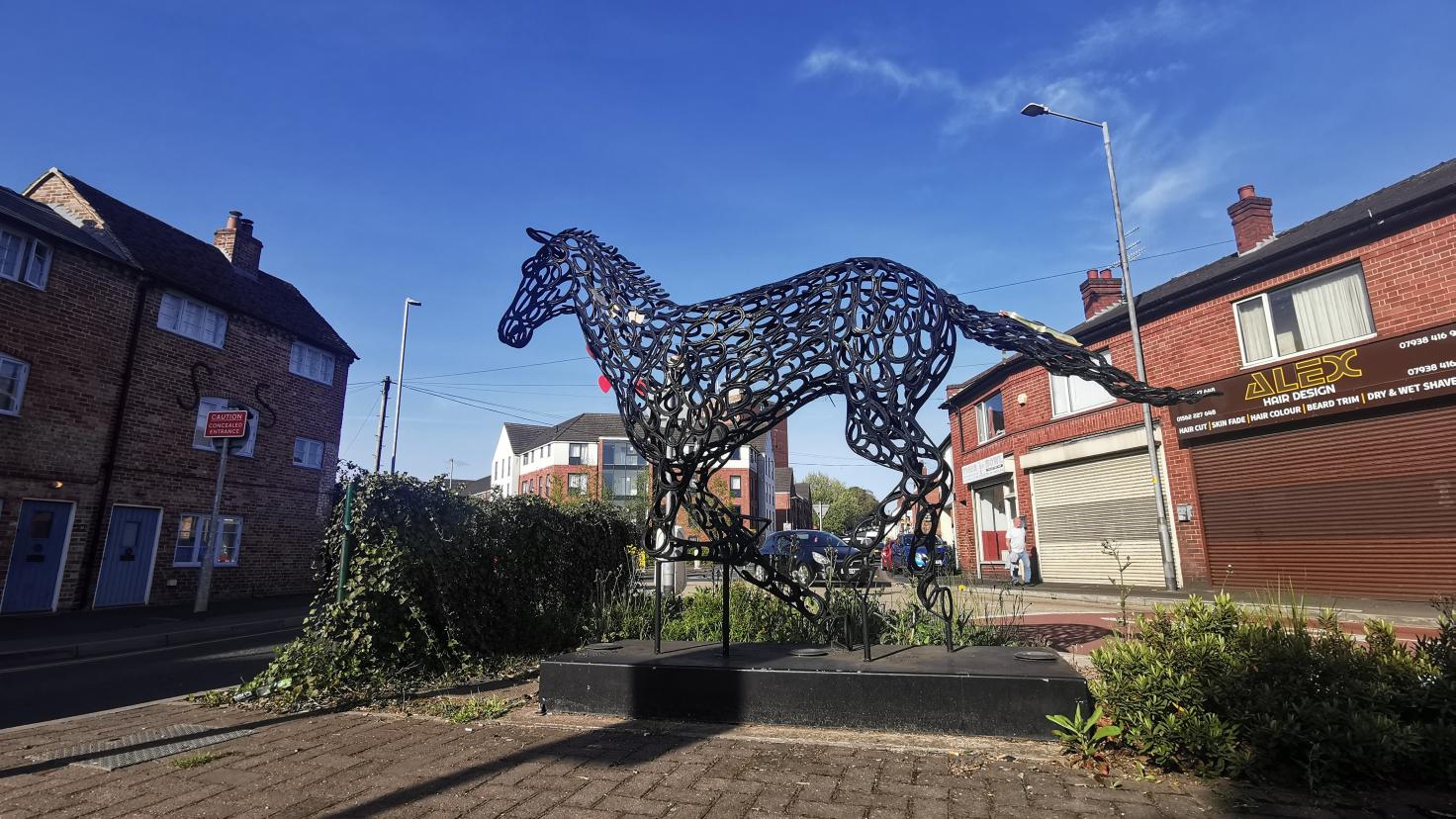 A black wire statue of a horse against a blue sky, in an area with buildings.