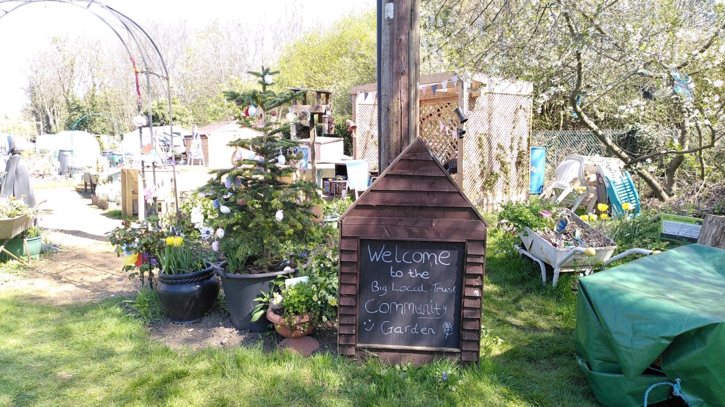 A garden with grass, plants and trees and a chalkboard sign reading 'Welcome'.