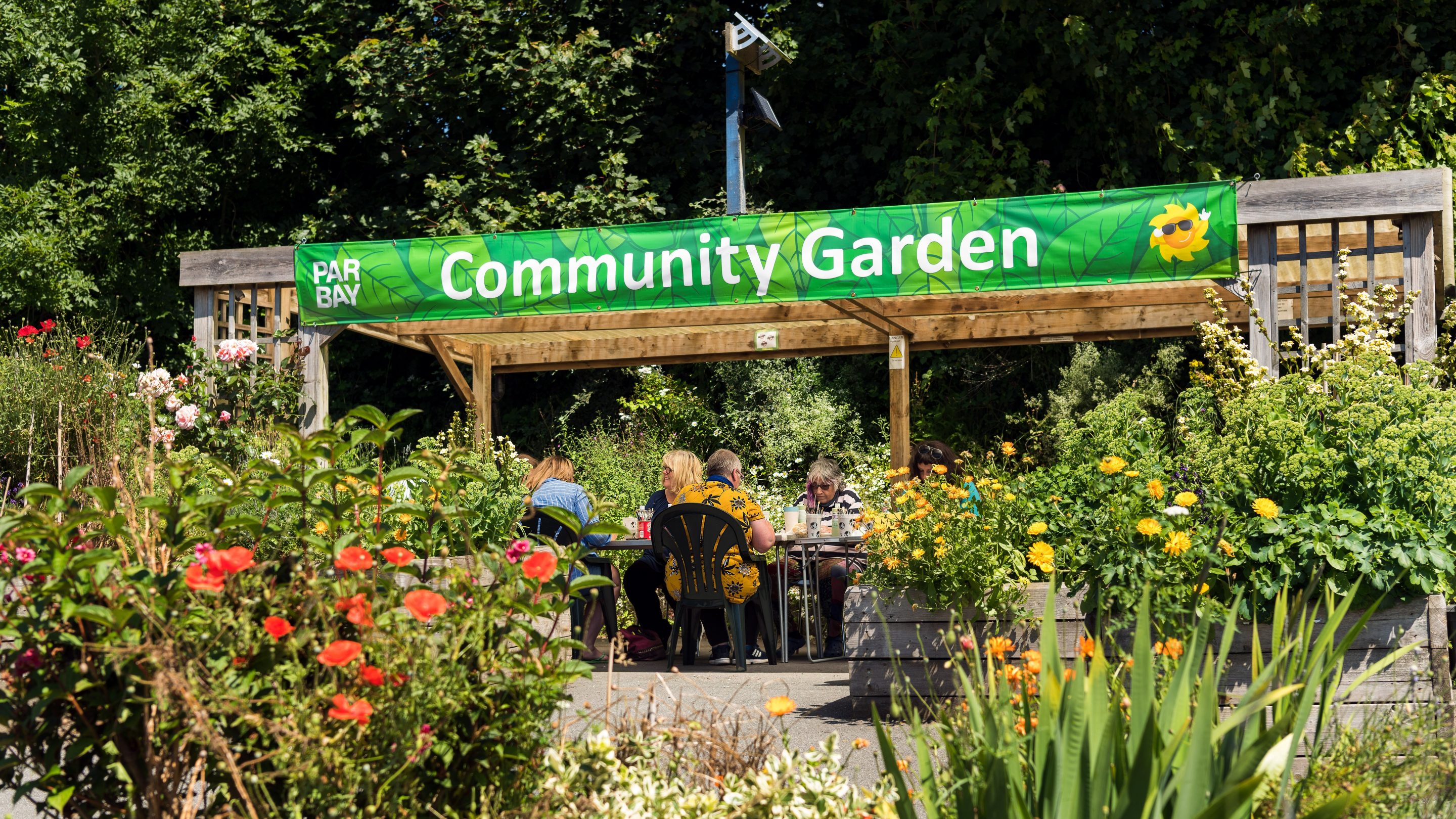 A group of people sitting around a table doing crafts under a pergola on a sunny day in the Par Bay Community Garden.