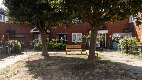 Wooden benches under a tree, with red-brick housing in the background.