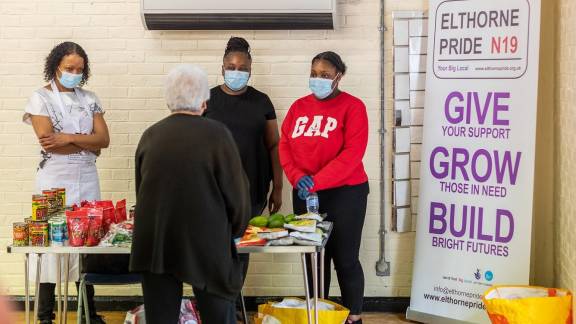 Three ladies behind a food stall, wearing face masks and serving an older lady who is perusing items.