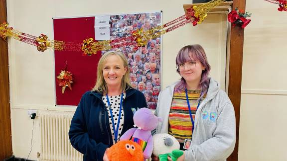 A woman with blonde hair and a young woman with pink hair stand next to each other, holding stuffed animals, and smiling at the camera. They have lanyards around their necks and colourful tops. Behind them is a yellow and red festive garland, set against a pale yellow brick wall.