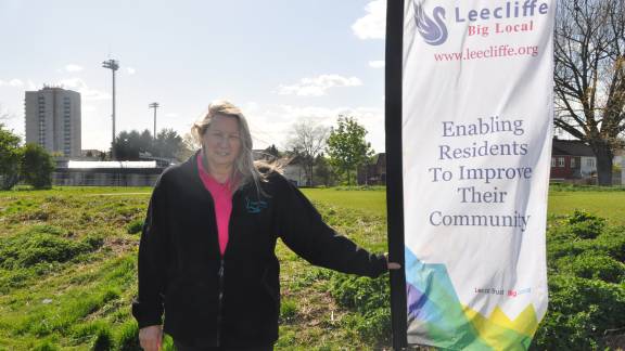 A woman with blonde hair, a pink polo shirt and a black fleece stands next to a flag that reads "Leecliffe Big Local. www.leecliffe.org Enabling residents to improve their community". She stands in a green field, with trees, houses and a tower block in the background.
