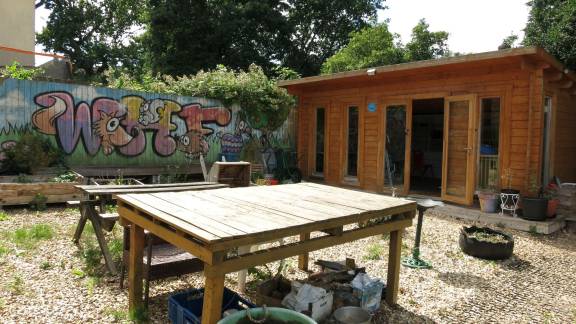 A wooden table in the sunshine in front of a wooden building, with a colourful spray-painted mural on a wall in the background.