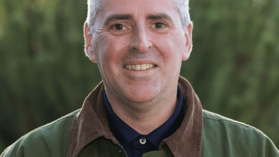 A man with grey hair and brown eyes wearing a green jacket with a brown collar and a navy shirt smiles at the camera. He stands against a blurred background of green bushes.